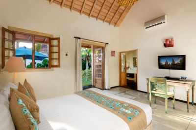 Spacious Balinese villa bedroom with wooden beamed ceiling, white king bed with teal accents, desk, TV, and tropical garden view through open shutters.