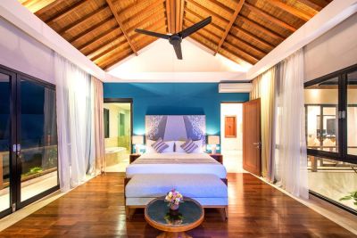 Spacious tropical bedroom with high wooden ceiling, king bed in blue tones, sheer-curtained balcony doors, and floral ottoman.
