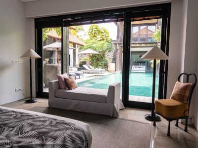Elegant Balinese villa bedroom with large sliding glass doors overlooking a turquoise infinity pool, tropical garden, and lounge area.