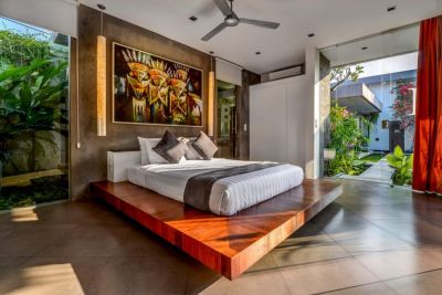 Luxurious Bali villa bedroom with wooden platform bed, tropical wall art, and lush garden view through glass doors.