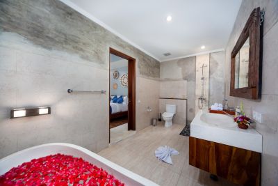 Luxurious Balinese villa bathroom with rose petal-filled bathtub, wooden vanity, toilet, and open bedroom view.