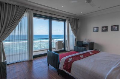Luxurious oceanfront bedroom with balcony view of turquoise waves, sheer curtains, king bed with red accents, and wooden floors.