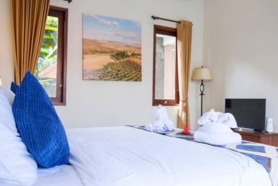 Cozy tropical bedroom with white bed, blue pillows, folded towels, landscape wall art, wooden accents, and lush outdoor views.