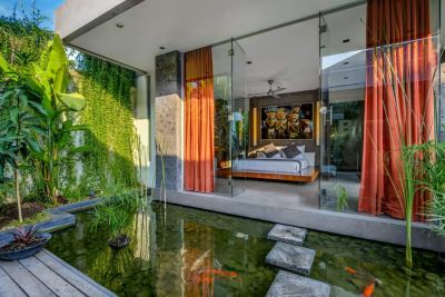 Modern tropical bedroom with floor-to-ceiling glass doors opening to lush greenery, koi pond, and orange curtains.