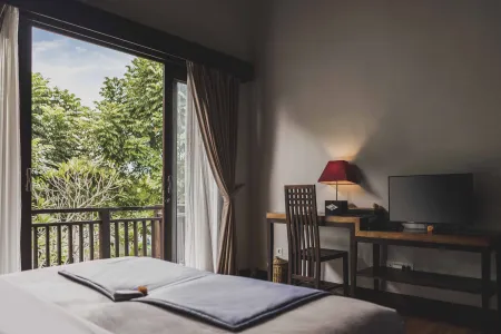 Serene tropical bedroom with open sliding doors to a leafy balcony view, wooden desk, chair, red lamp, and neatly made bed.
