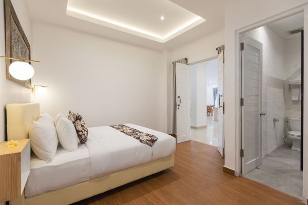 Cozy modern Bali bedroom with white walls, wooden floor, neatly made bed, wall lamp, and open door to adjacent bathroom.