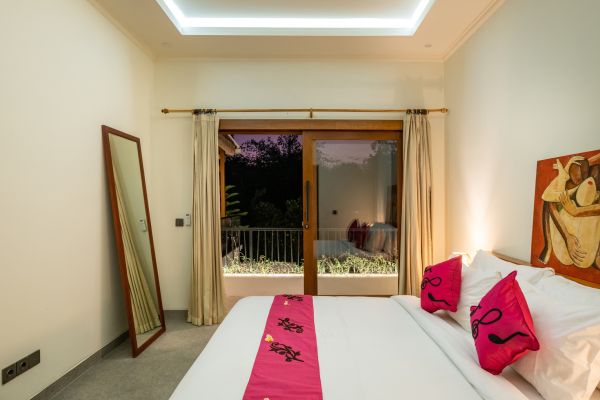 Serene tropical bedroom with white bed, pink pillows, wooden balcony doors to lush garden view at dusk.