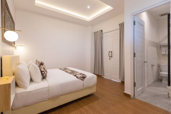 Spacious modern bedroom with white walls, king bed, wooden floors, and open door to en-suite bathroom.