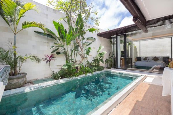 Private infinity pool in lush tropical Bali villa garden, with palm trees and open glass doors to bedroom.