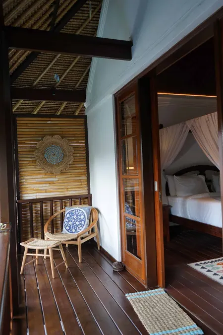 Open wooden balcony in a tropical villa, with rattan chair, woven bamboo wall art, and sliding doors to a canopy bed bedroom.