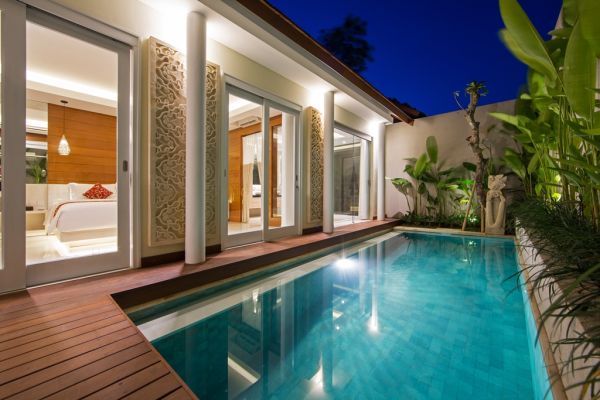 Luxurious Balinese villa bedroom with open sliding doors overlooking a lit private pool at night, surrounded by tropical plants.