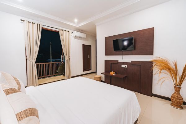 Cozy modern hotel bedroom with white linens on a king bed, wall-mounted TV, air conditioner, potted plant, and open balcony doors revealing a nighttime city view.