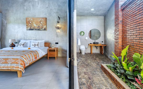 Split-view of a luxurious Balinese villa bedroom with wooden bed, orange batik linens, and wall art, connected to a modern bathroom featuring a round mirror, wooden vanity, and tropical plants.