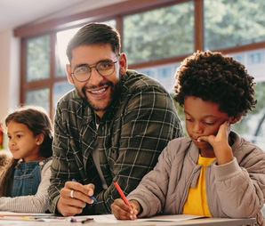 Primary,Educator,Smiling,At,The,Camera,In,A,Classroom.,Teacher Primary educator smiling at the camera in a classroom. Teacher teaching a group of children in a school. Man giving an art lesson in an early child development centre.