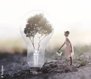 Eine riesige Glühbirne steht auf einem Feld. Statt des Glühdrahts sind in ihrem Inneren Äste zu einem Baum verwachsen. Eine Frau steht vor der Glühbirne und gießt sie mit einer Kanne.