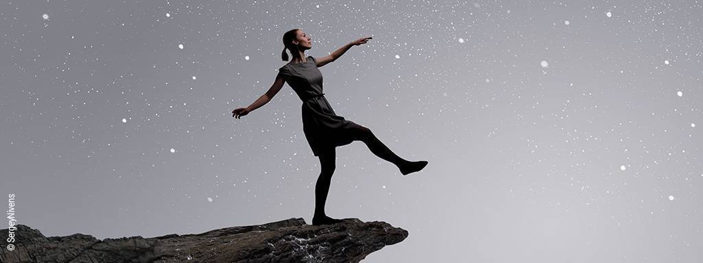 Eine Frau steht mit ausgestrecktem Fuß an einer Klippe vor Sternenhimmel.