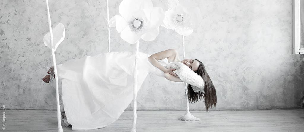 Foto: Eine Person im weißen Kleid schwebt liegend über Holzdielen inmitten überdimensionierter Papierblumen. Der Kopf liegt auf einem Seitenschläferkissen.