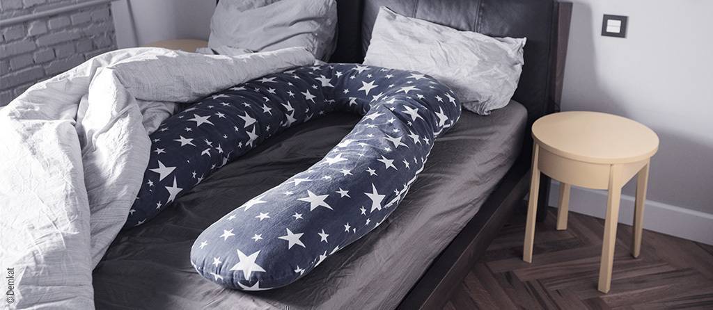 Foto: Ein U-förmiges Seitenschläferkissen mit weißen Sternen auf blauem Grund liegt auf einer Bettseite im Doppelbett.