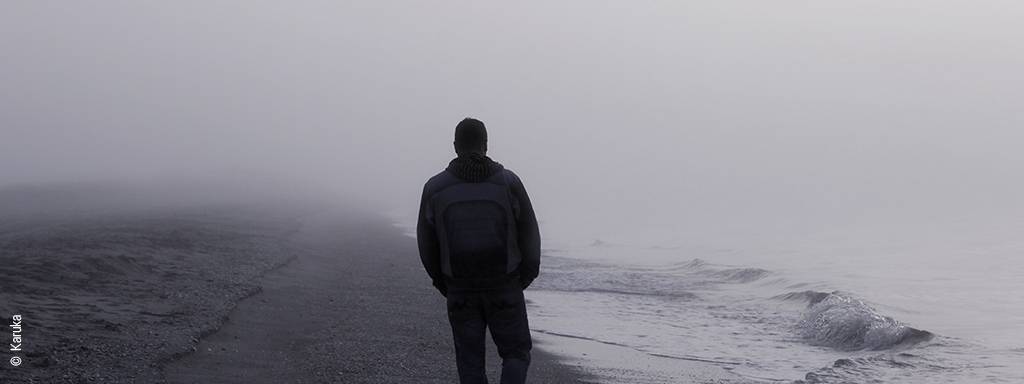 Foto: Eine Person geht einen leeren Strand im grauen Nebel entlang. Sie trägt einen Rucksack auf dem Rücken und ist von hinten zu sehen.