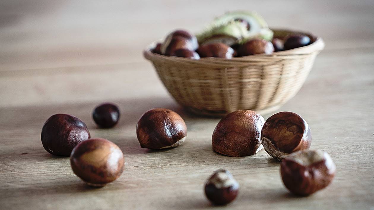 Foto: Ein Korb mit Kastanien steht auf einem Holztisch, davor liegen einige einzelne Kastanien.