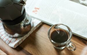 Foto: Ein Holztisch in Vogelperspektive. Darauf eine Kaffeekanne, ein Glas mit Espresso und die Morgenlektüre.