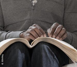 Foto: Ein aufgeschlagenes Buch auf dem Schoss einer blinden Person. Die Hände lesen Brailleschrift.