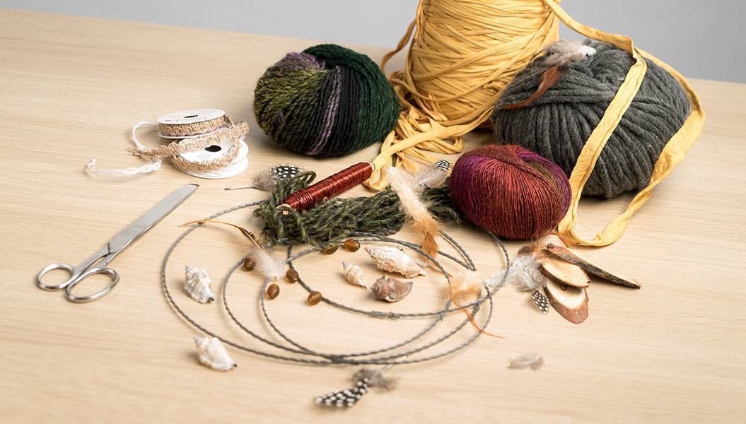Foto: Bastelmaterial für Traumfänger auf einem Holztisch: Verschiedene Arten von Wolle, Garn und Bändern, Draht, Federn, Holzscheiben, Muscheln und eine Schere.