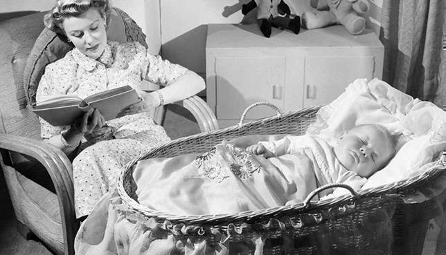 Foto: Ein Baby liegt in einer Wiege. Im Hintergrund sitzt die Mutter und liest aus einem Buch vor.