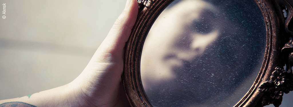 Eine Hand hält einen verzierten Spiegel. Darin spiegelt sich schemenhaft ein Gesicht.
