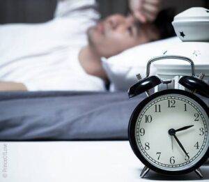 Foto: ein Wecker, im Hintergrund eine wachliegende Person im Bett