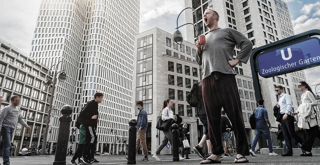 Foto: Vor dem U-Bahneingang einer Großstadt steht eine Person herzhaft gähnend mit Kaffeetasse und Pyjama in einer Menschenmenge.