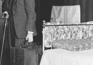 Historische Schwarz-Weiß-Fotografie: Ein Herr mit Gehstock, Koffer steht zum Gehen gewandt im Anzug vor einem gemachten Bett.