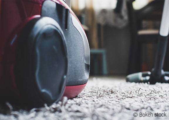 Foto: Auf einem grauen Teppich steht ein roter Staubsauger. Im Hintergrund ist eine Wohnzimmereinrichtung zu erkennen.