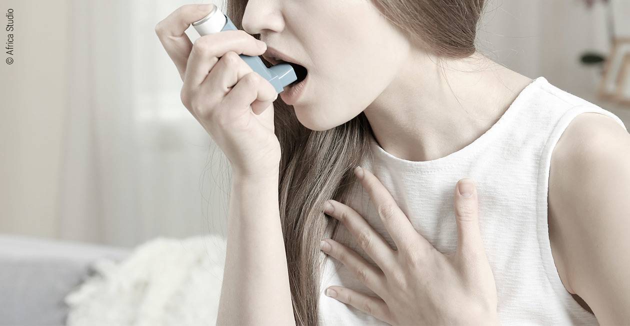 Foto: Eine junge Frau hält sich mit einer Hand den Brustkorb. Mit der anderen Hand führt sie einen Asthma-Inhalator zum Mund.