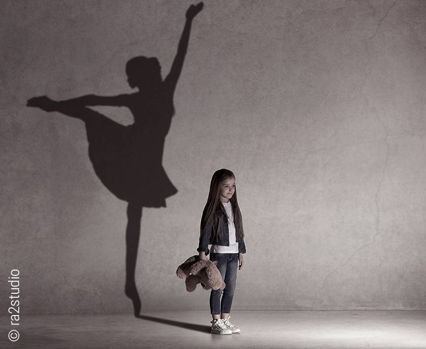 Foto: Ein kleines Mädchen mit Teddybären in der Hand steht vor einer Wand. Ihr Schatten zeigt eine tanzende Ballerina.