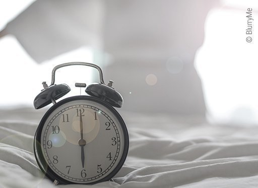 Foto: Auf einem Bett mit weißer Bettwäsche steht eine Zeigeruhr. Die Uhr zeigt sechs Uhr. Im Hintergrund streckt sich eine Person vor einem von Licht erhellten Fenster.