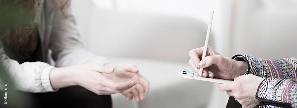 Foto: Zwei Personen sitzen sich bei einer Therapiesitzung gegenüber; eine Person hält Block und Stift in der Hand.