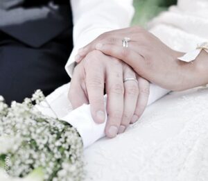 Foto: Traumdeutung Hochzeit / Hochzeitskleid – die Hände eines Brautpaares liegen übereinander auf einem Blumenstrauß.