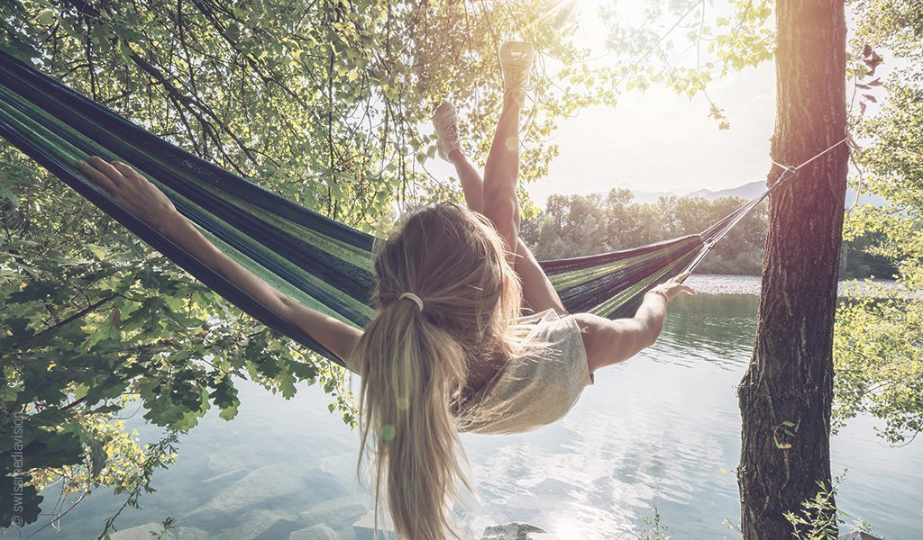 Foto: eine Hängematte hängt vor einem See. Die Person in der Hängematte scheint stark zu schaukeln oder herauszufallen: die Füße sind in der Luft der Oberkörper auf der anderen Seite der Hängematte neigt sich dem Boden zu