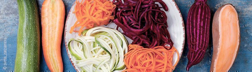 Foto: Zucchini, Möhre, rote Beete und Süßkartoffeln liegen auf einem Tisch - in der Mitte ein Teller auf dem spagetthiförmige Streifen aus ihnen geschnitten und drapiert wurden.