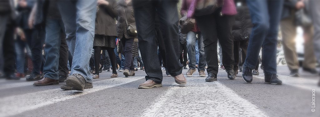 Foto: Auf einer asphaltierten Straße gehen viele Menschen. Es sind nur die Füße und Beine zu sehen. 