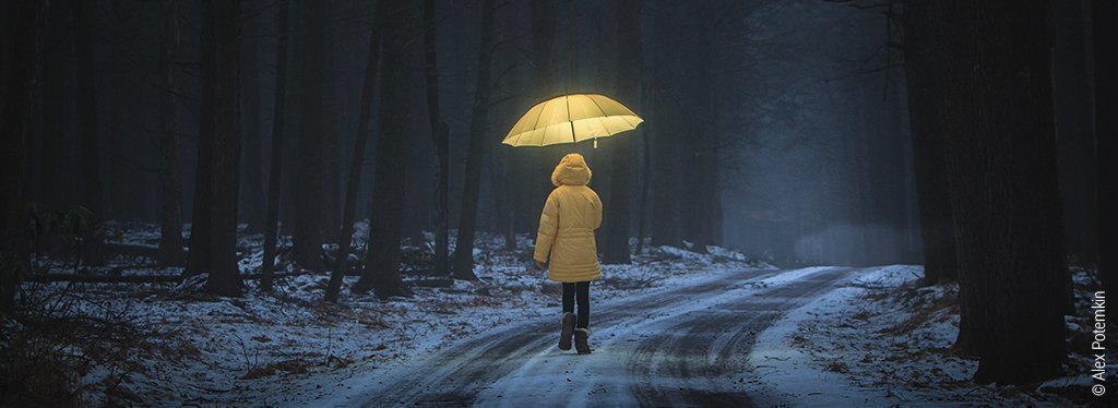 Foto: Ein Kind mit gelbem Mantel und gelb erleuchtetem Regenschirm geht einen dunklen Waldweg entlang. Eine dünne Schicht Schnee liegt auf dem Boden.