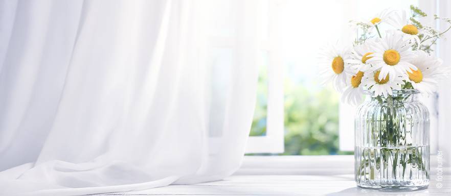 Foto: Eine Vase mit Blumen steht auf einem Fensterbrett, das Fenster ist leicht geöffnet und die Sonne scheint.