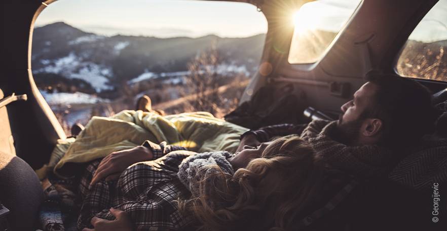 Foto: Ein Pärchen liegt im Auto mit Blick auf ein Bergpanorama. Die Sonne strahlt über einen Gipfel.