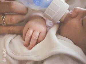 Foto: Ein Baby liegt mit geschlossenen Augen auf dem Rücken und wird von Hand mit einer Flasche gefüttert.