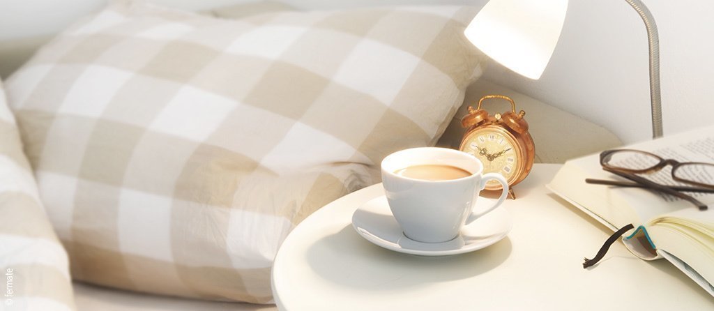 Foto: In einem Bett liegt beige karierte Bettwäsche. Davor steht ein runder Nachttisch auf dem Kaffeetasse, Wecker, Lampe, Buch und Lesebrille platziert sind.