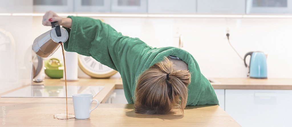  Foto: In der Küche sitzt eine Person am Tisch, den Kopf auf der Tischplatte. Die rechte Hand ist erhoben und hält eine Kaffeekanne, aus der der Kaffee neben eine darunter platzierte Tasse fließt.