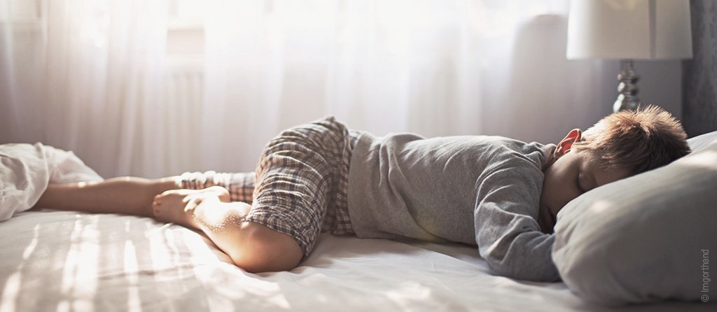 Foto: Ein Kind liegt auf dem Bauch im Bett und schläft ohne Bettdecke. Die Sonne scheint durchs Fenster auf die Matratze.