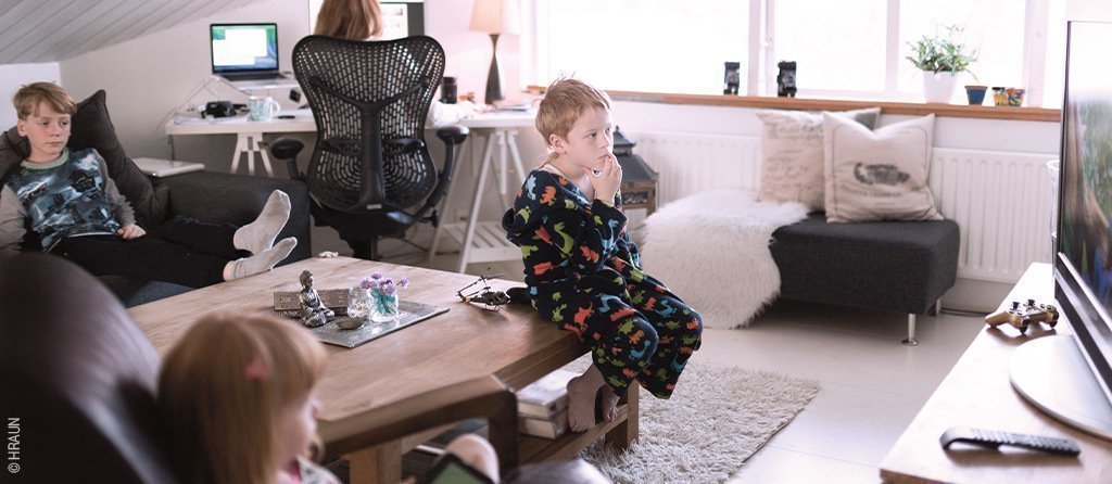 Foto: Drei Kinder schauen im Wohnzimmer Fernsehen, im Hintergrund arbeitet eine Frau am Computer