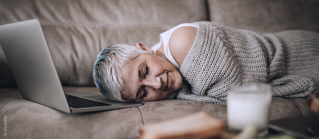 Foto: Eine Frau liegt auf dem Bauch schlafend auf einem Sofa, vor ihr steht ein Laptop.